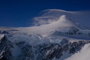 A snow-covered mountain peak rises sharply under a deep blue sky, with wispy clouds trailing from the summit and steep, rugged slopes below covered in ice and snow.