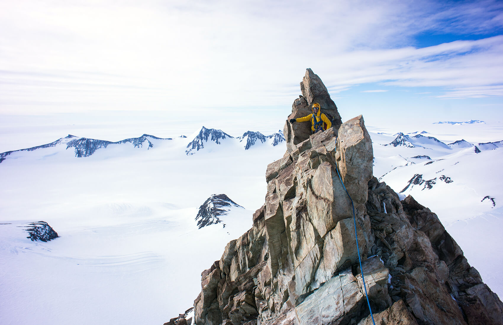 A climber in yellow gear stands on a jagged rocky peak in Antarctica, surrounded by snow-covered mountains and a bright blue sky. Ropes along the rocks highlight the challenging ascent with Antarctic Logistics and Expeditions support.