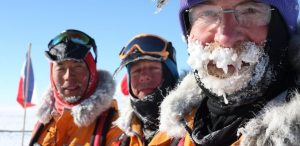 Three people dressed in heavy winter gear stand outdoors in snowy conditions. The person on the right has ice and frost on their beard and mustache. A Russian flag is visible in the background against a clear blue sky.