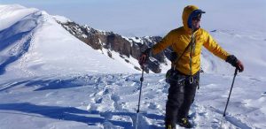 A person in a yellow jacket and snow gear stands on a snowy mountain ridge, holding trekking poles, with snow-covered peaks and a rocky cliff in the background under a clear sky.