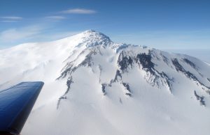 A snow-covered mountain with dark rocky patches in Antarctica is seen from an airplane operated by Antarctic Logistics and Expeditions, with the wing visible in the foreground under a clear blue sky.