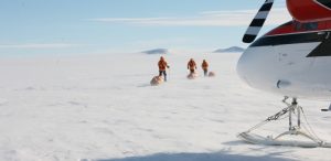 Four people in orange jackets pull sleds across a snowy, flat landscape. A ski-equipped aircraft is parked in the foreground. Distant hills are visible under a clear sky.