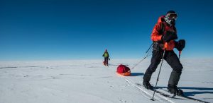 Two people in winter gear ski across a vast, snowy landscape under a clear blue sky, pulling sleds loaded with supplies behind them.