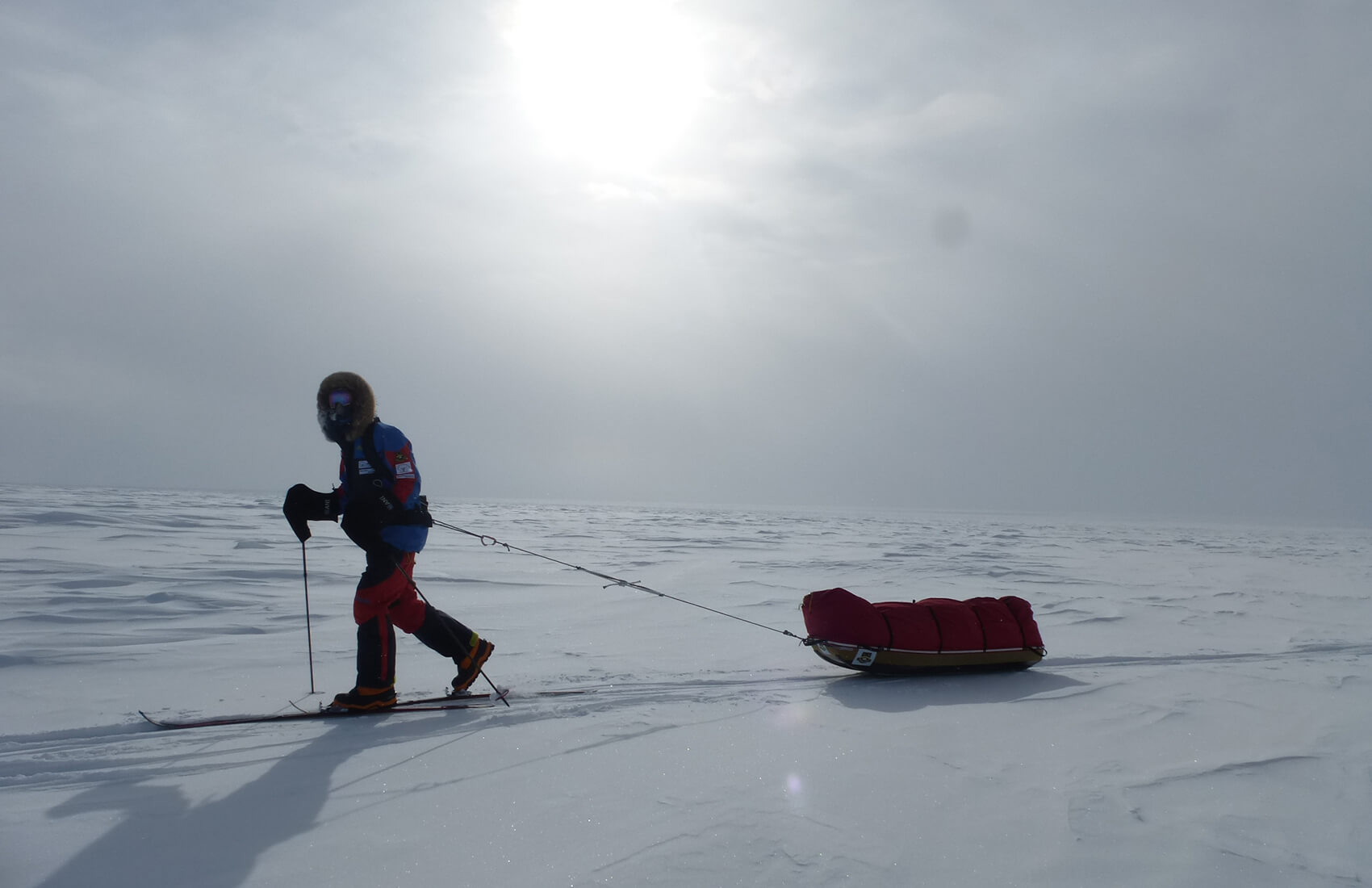 A person in winter gear pulls a red sled across a vast, snowy Antarctic landscape under a cloudy sky, with the sun faintly shining through—capturing the spirit of Antarctica and Antarctic Logistics and Expeditions.