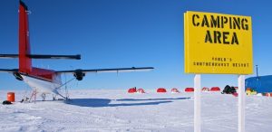A small red and white plane is parked on snowy ground near a yellow sign that reads CAMPING AREA: WORLDS SOUTHERNMOST RESORT. Red tents are set up in the background under a clear blue sky.