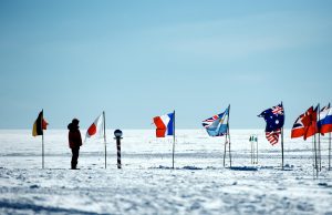 A person in a red parka stands on snowy ground in Antarctica near a striped marker surrounded by several international flags, including Germany, Japan, France, the UK, Australia, and Russia—an adventure with Antarctic Logistics and Expeditions.