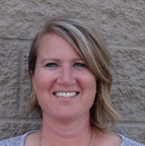 A woman with shoulder-length blonde hair, wearing hoop earrings and a gray top, smiles in front of a textured beige brick wall, radiating the adventurous spirit often found among Antarctic Logistics and Expeditions teams.