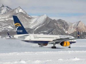 A white and blue Icelandair airplane with yellow engines, operated by Antarctic Logistics and Expeditions, is parked on a snowy runway, surrounded by snow-covered mountains under a clear sky.