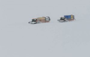 Two snow vehicles from Antarctic Logistics and Expeditions pull sleds loaded with cargo containers and supplies across the vast, snowy expanse of Antarctica, barely visible through the blowing snow.