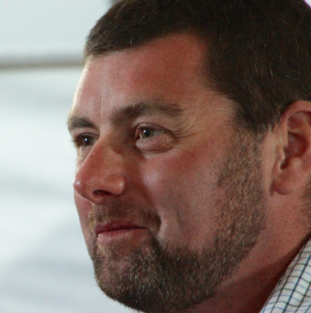A close-up of a man with short brown hair and a beard, wearing a plaid shirt, looking slightly to the left with a gentle smile and soft lighting in the background—echoing the warmth found amid Antarctic Logistics and Expeditions.