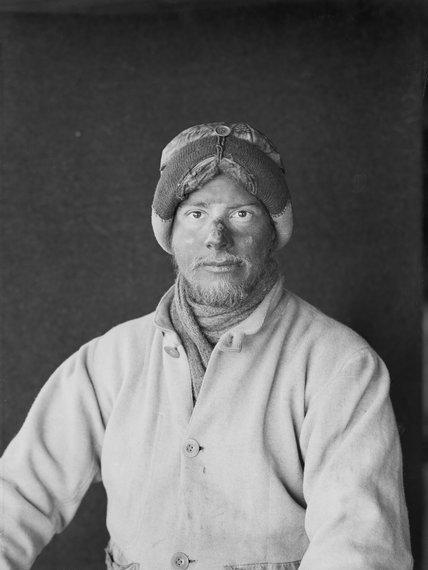 A black-and-white portrait of a bearded man in a heavy, buttoned coat and thick hat with ear flaps, gazing at the camera—capturing the spirit of Antarctic Logistics and Expeditions against a plain dark background.