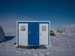 A small blue and white building with two doors stands on a snowy Antarctic landscape under a clear blue sky. A yellow snowmobile and large white tents, part of Antarctic Logistics and Expeditions, are visible in the background.