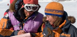 Two people dressed in cold-weather gear stand on snow in Antarctica, examining a pink sheet of paper. Snowy mountains and others are visible in the background. Both wear jackets, hats, and goggles—part of an Antarctic Logistics and Expeditions outing.