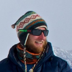 A person with a beard wearing a colorful knit hat, dark sunglasses, a scarf, and a blue jacket, smiling outdoors in Antarctica with a snowy mountain in the background—perfect for an Antarctic Logistics and Expeditions adventure.