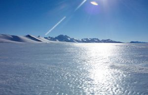 A bright sun shines over an icy, snow-covered landscape in Antarctica, with distant mountain peaks under a clear blue sky. Sunlight reflects off the frozen surface near the South Pole, creating a glittering effect.