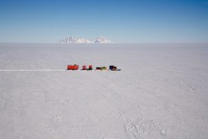 A line of vehicles and small buildings travels across a vast, snowy landscape in Antarctica under a clear blue sky, with distant mountains on the horizon—typical of Antarctic Logistics and Expeditions en route to the South Pole.