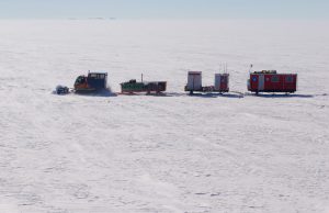 A snow tractor pulls three sleds loaded with equipment and a red mobile shelter across a vast, snowy, and flat Antarctic landscape under a clear sky, supporting Antarctic Logistics and Expeditions near the South Pole.