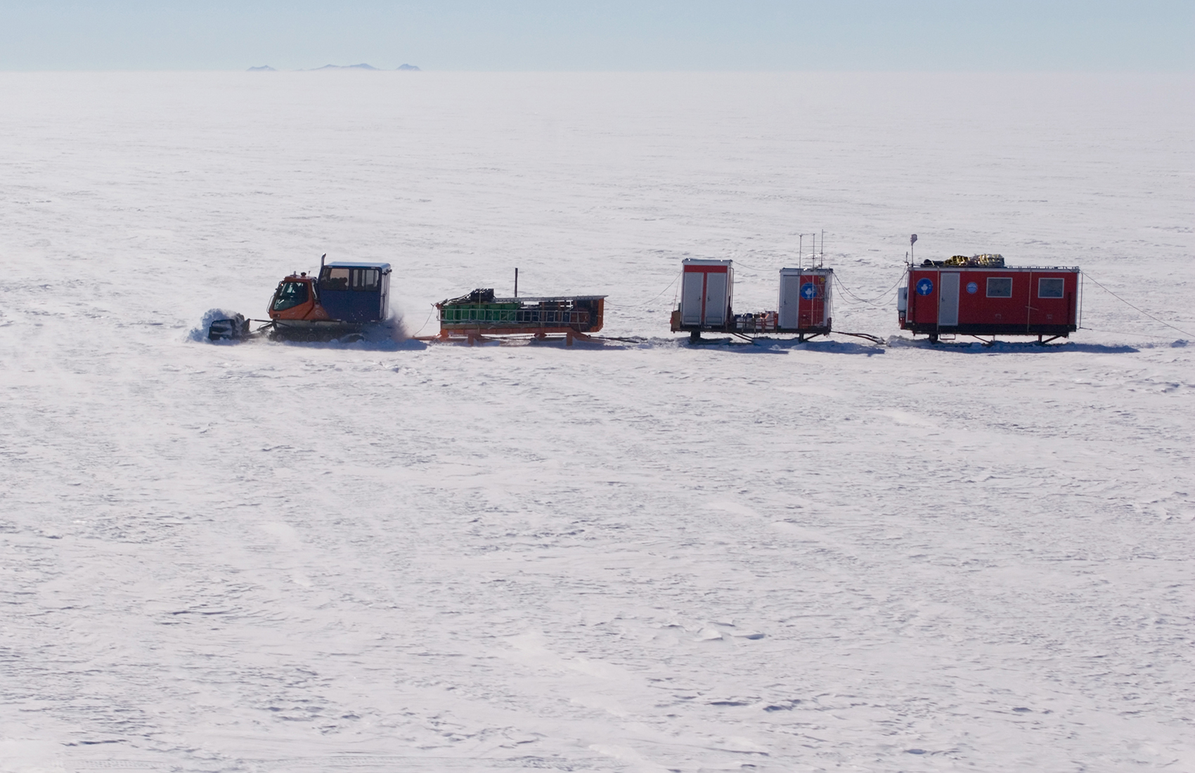 A snow tractor pulls three sleds loaded with equipment and a red mobile shelter across a vast, snowy, and flat Antarctic landscape under a clear sky, supporting Antarctic Logistics and Expeditions near the South Pole.