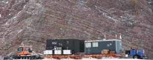 In Antarctica, a snow tractor pulls two portable research labs and supplies on sleds across a snowy, rocky landscape with steep, layered cliffs in the background, showcasing the vital role of Antarctic Logistics and Expeditions.