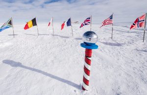 A shiny silver globe on a red-and-white striped pole stands in snowy terrain at Antarctica's South Pole, surrounded by international flags under a cloudy sky—a landmark often visited with Antarctic Logistics and Expeditions.