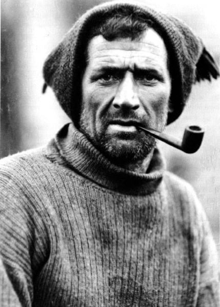 Black-and-white portrait of a rugged man in a knit sweater and ear-flap hat, gazing seriously at the camera with a pipe in his mouth—reminiscent of early Antarctic Logistics and Expeditions explorers.
