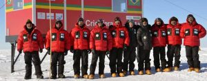 Ten people in red jackets and snow boots stand side by side on snow in front of a red building with Brazilian flags and logos, likely at an Antarctic Logistics and Expeditions site in Antarctica.