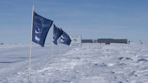 Blue flags stand in snow leading to a sign and a large building in the distance under a clear South Pole sky. The scene is cold and remote, with tire tracks visible—hallmarks of Antarctic Logistics and Expeditions operations.