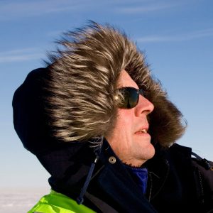 A person in a fur-lined hooded parka and sunglasses looks upward against a clear blue sky, capturing the spirit of Antarctica and Antarctic Logistics and Expeditions in a cold, sunny environment.