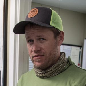 A man wearing a green shirt, camouflage neck gaiter, and a green and black cap stands indoors, possibly at an Antarctic Logistics and Expeditions facility. Papers and a bulletin board are visible in the background as he looks at the camera with a neutral expression.