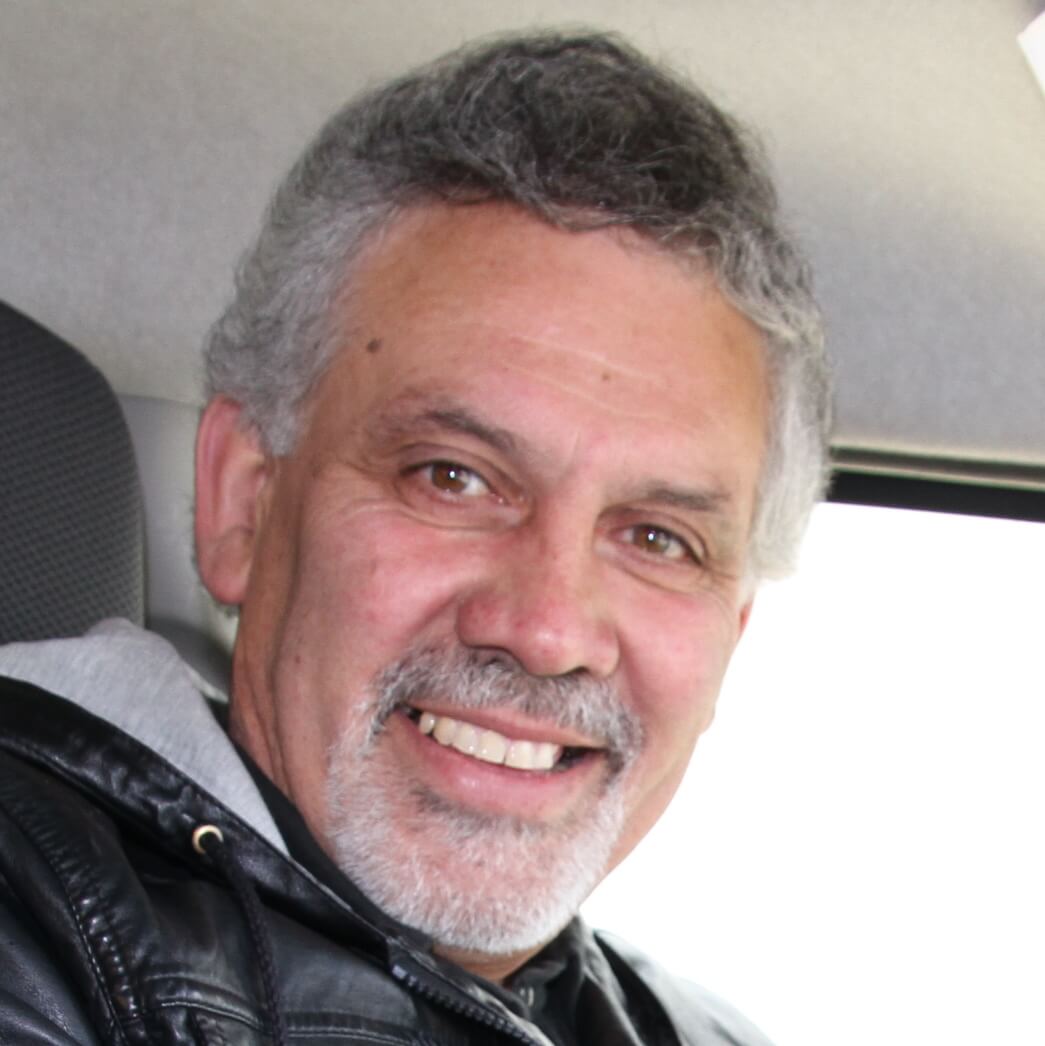 A middle-aged man with short curly gray hair and a beard smiles at the camera while sitting indoors, wearing a black leather jacket and a gray hoodie—perhaps reminiscing about his recent Antarctic Logistics and Expeditions adventure.