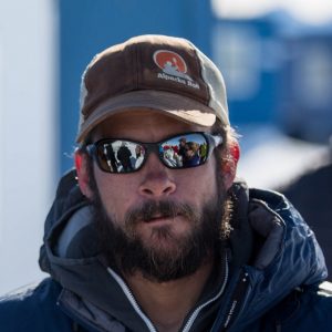 A man with a beard, wearing a brown Alpacka Raft cap, dark sunglasses, and a blue jacket stands outside in Antarctica. Reflections of people and snowy surroundings hint at his connection to Antarctic Logistics and Expeditions.