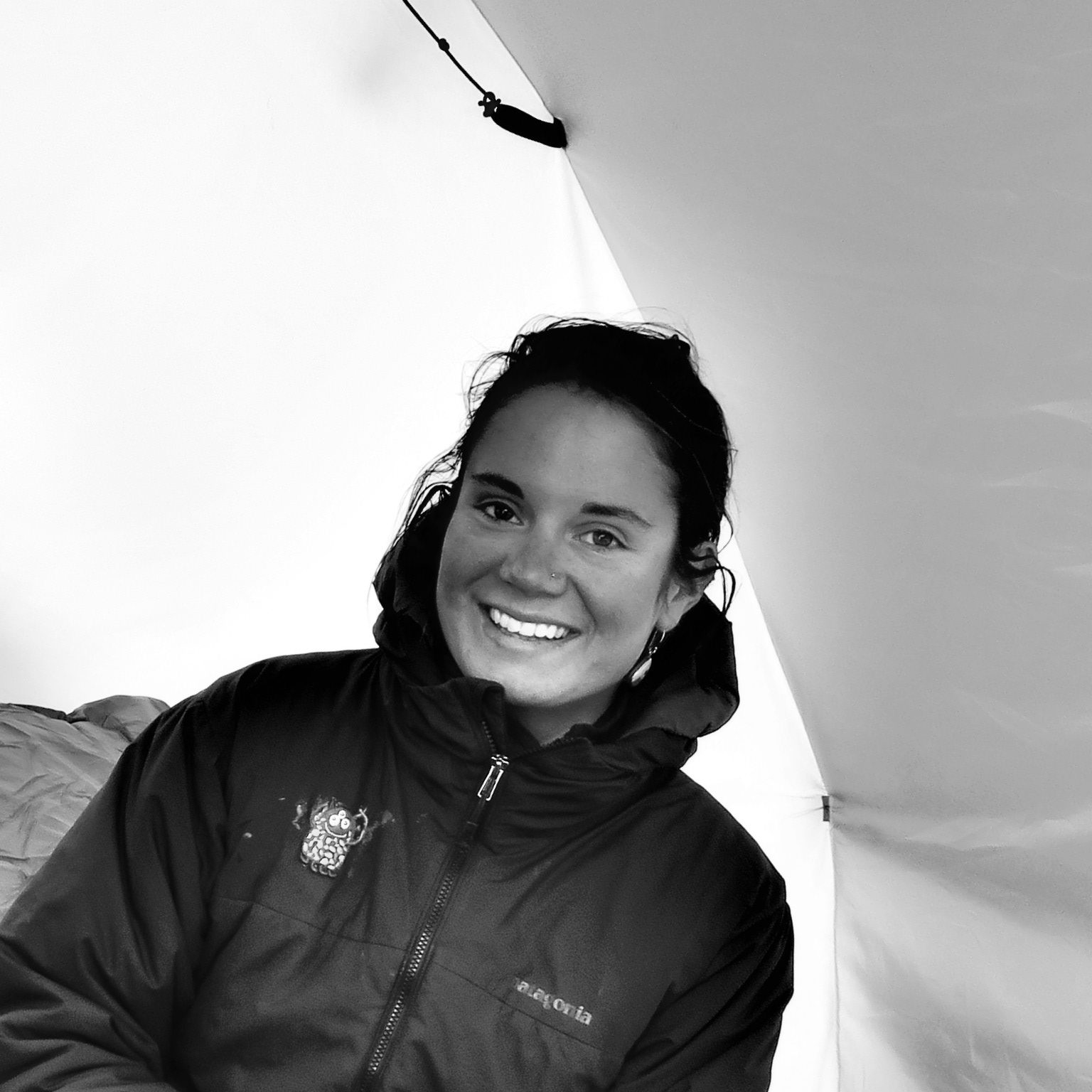 A woman smiles inside a tent, wearing a hooded jacket. The black and white image captures the interior fabric—echoing the spirit of Antarctic Logistics and Expeditions in the heart of Antarctica.