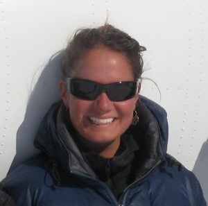 A woman wearing dark sunglasses and a blue jacket smiles at the camera, ready for her Antarctic adventure. She is standing in front of a white background, echoing the icy landscapes supported by Antarctic Logistics and Expeditions.