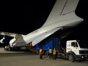Three workers in safety vests oversee cargo being loaded from a truck into the open rear of a large Antarctic Logistics and Expeditions airplane at night on an airport tarmac.