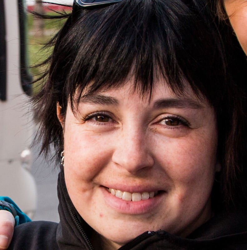 A woman with short dark hair smiles at the camera, wearing a black jacket and sunglasses on her head. The background is slightly blurred, hinting at an outdoor Antarctica setting during an Antarctic Logistics and Expeditions adventure.
