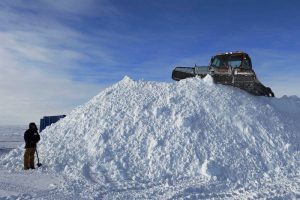 A person stands with a shovel at the base of a large snow pile while a bulldozer pushes snow at the top, under a mostly cloudy sky in Antarctica, showcasing the efforts of Antarctic Logistics and Expeditions.