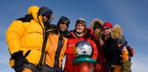 Five people dressed in heavy winter gear stand close together smiling beside a shiny silver sphere atop a red and green pole, with Antarctica’s blue sky in the background—a memorable moment supported by Antarctic Logistics and Expeditions.