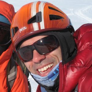 A person wearing an orange helmet, dark sunglasses, and red jacket smiles outdoors in snowy Antarctica, possibly part of an Antarctic Logistics and Expeditions adventure.