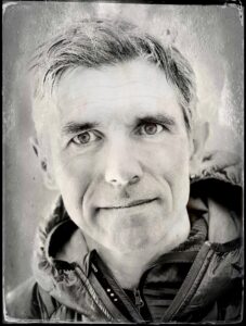 Black and white close-up portrait of a middle-aged man with short hair, wearing a zip-up jacket, looking directly at the camera with a neutral expression. The blurred background hints at his experience with Antarctic Logistics and Expeditions.