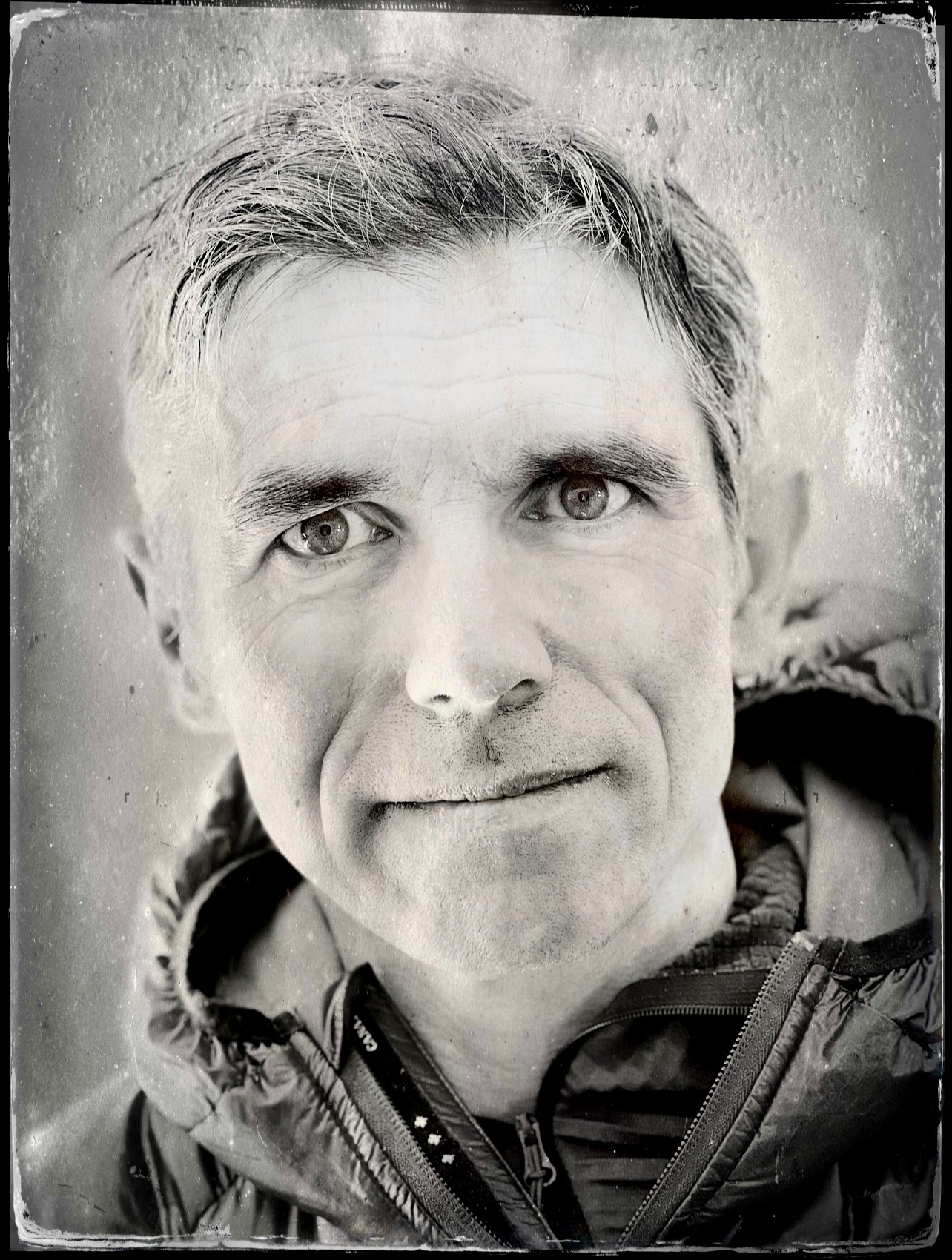 Black and white close-up portrait of a middle-aged man with short hair, wearing a zip-up jacket, looking directly at the camera with a neutral expression. The blurred background hints at his experience with Antarctic Logistics and Expeditions.