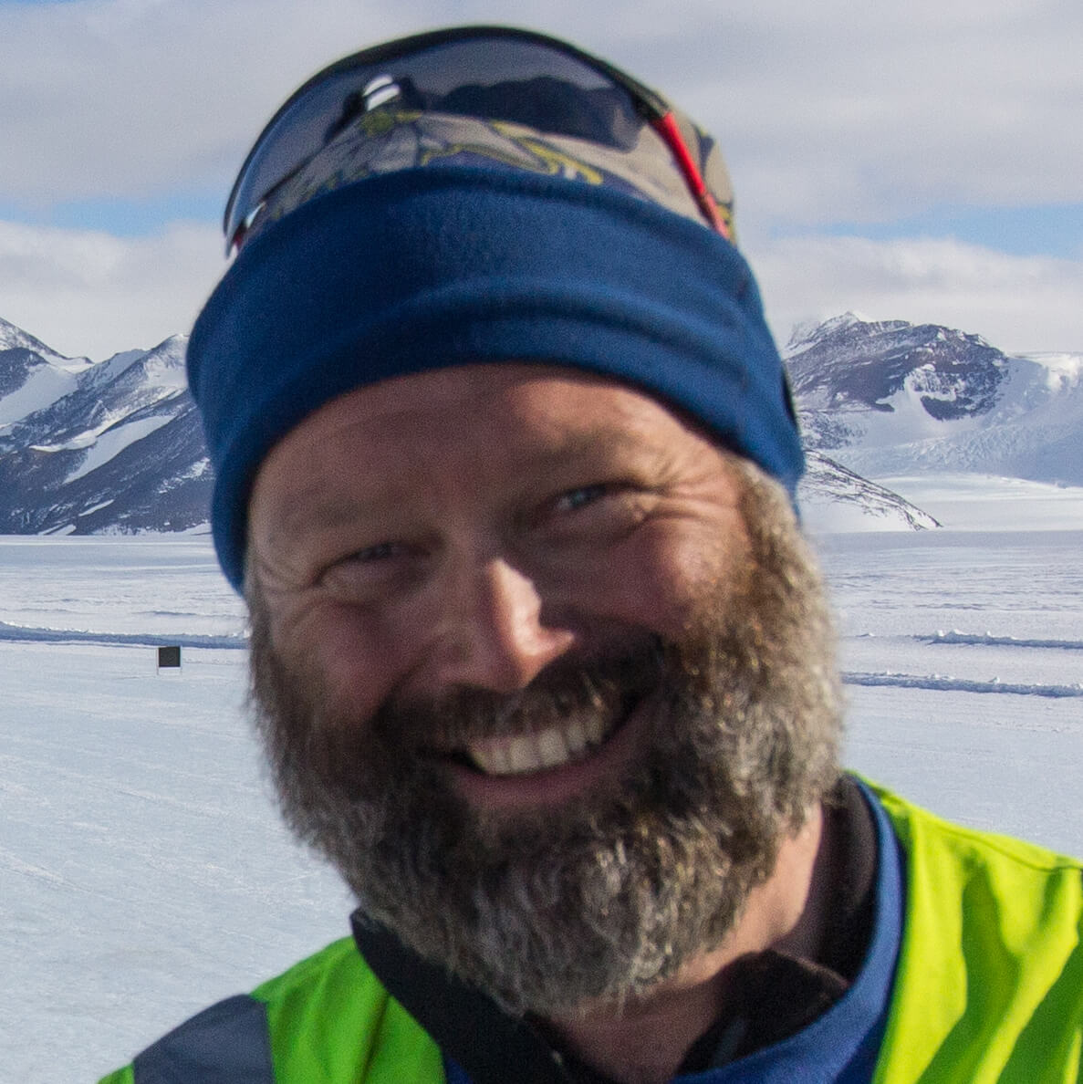 A bearded man in a blue hat and sunglasses smiles outdoors in a snowy, mountainous landscape with bright skies, reminiscent of an Antarctica adventure with Antarctic Logistics and Expeditions.