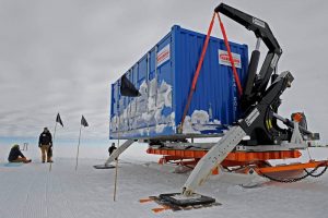 A blue shipping container secured on an orange sled is lifted by a black crane in Antarctica's snowy landscape. Three people from Antarctic Logistics and Expeditions are nearby, with black flags marking the area and snow clinging to the container’s side.