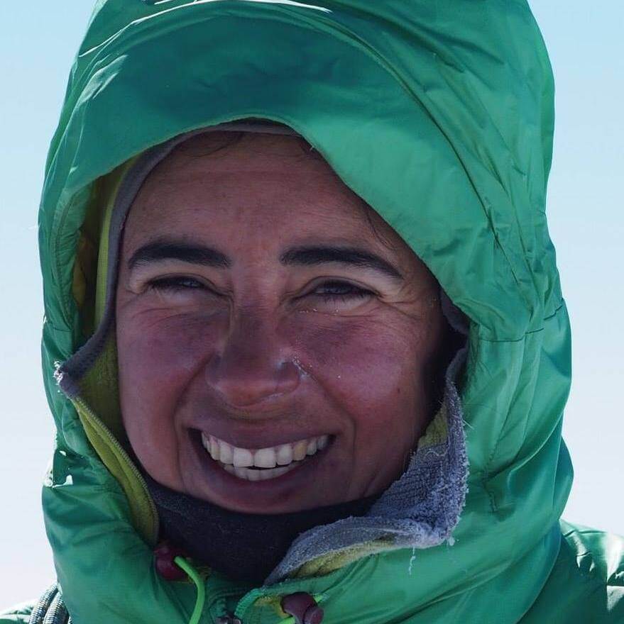 A person smiling broadly in Antarctica, wearing a green hooded winter jacket and outdoor gear from Antarctic Logistics and Expeditions, with frost on their collar and sunlight warming their face.