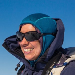 A person smiling outdoors in Antarctica, wearing sunglasses, a blue hooded jacket with several layers, and carrying a backpack. The clear blue sky in the background hints at an adventure with Antarctic Logistics and Expeditions.