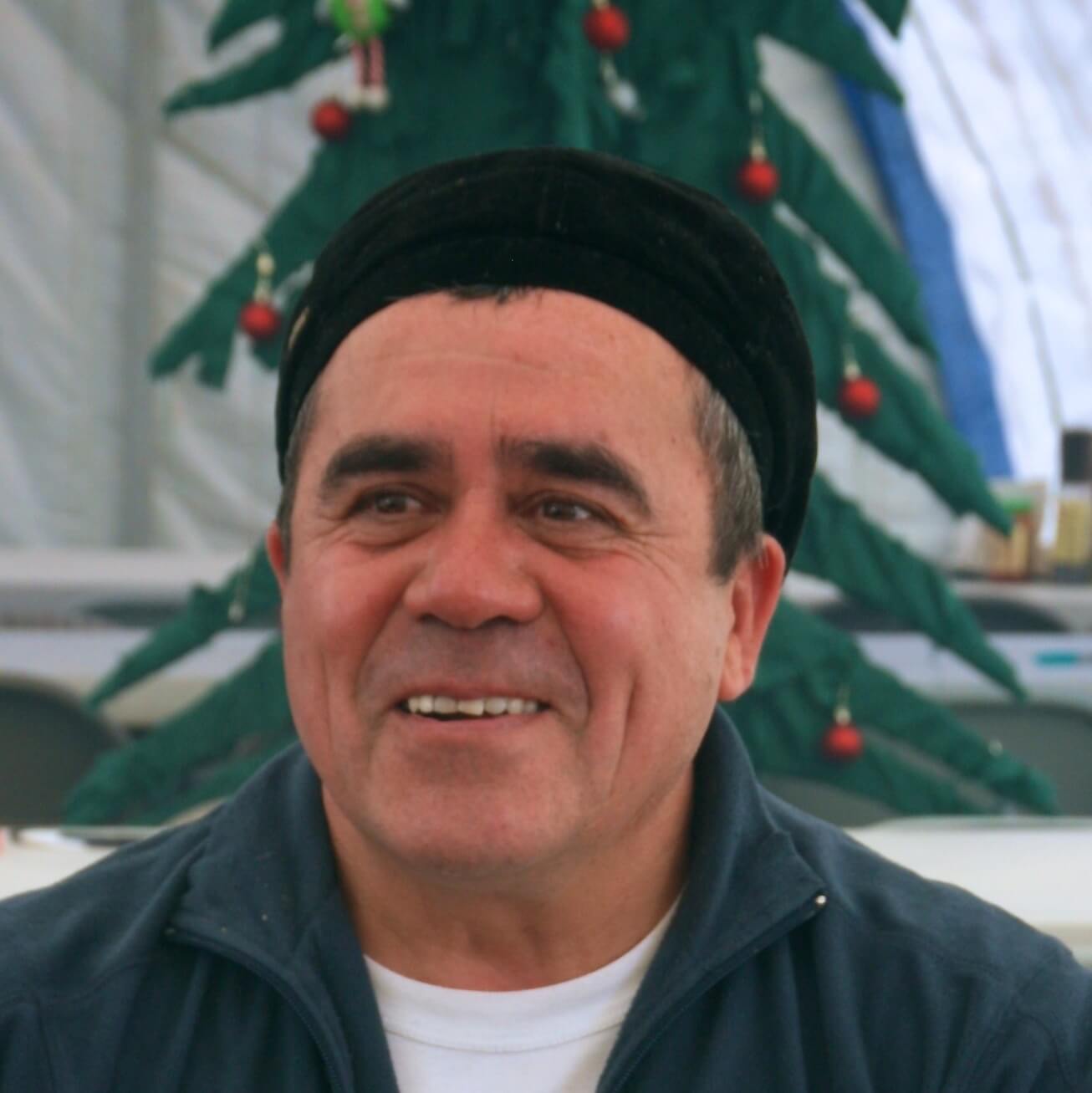 A middle-aged man wearing a dark cap and a zip-up jacket smiles warmly. In the background, there is a decorated Christmas tree with ornaments—perhaps bringing holiday cheer to an Antarctic Logistics and Expeditions camp.