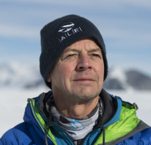 A man wearing a dark beanie and blue-green winter jacket stands outdoors in snowy, mountainous terrain near the South Pole, looking into the distance under bright daylight.