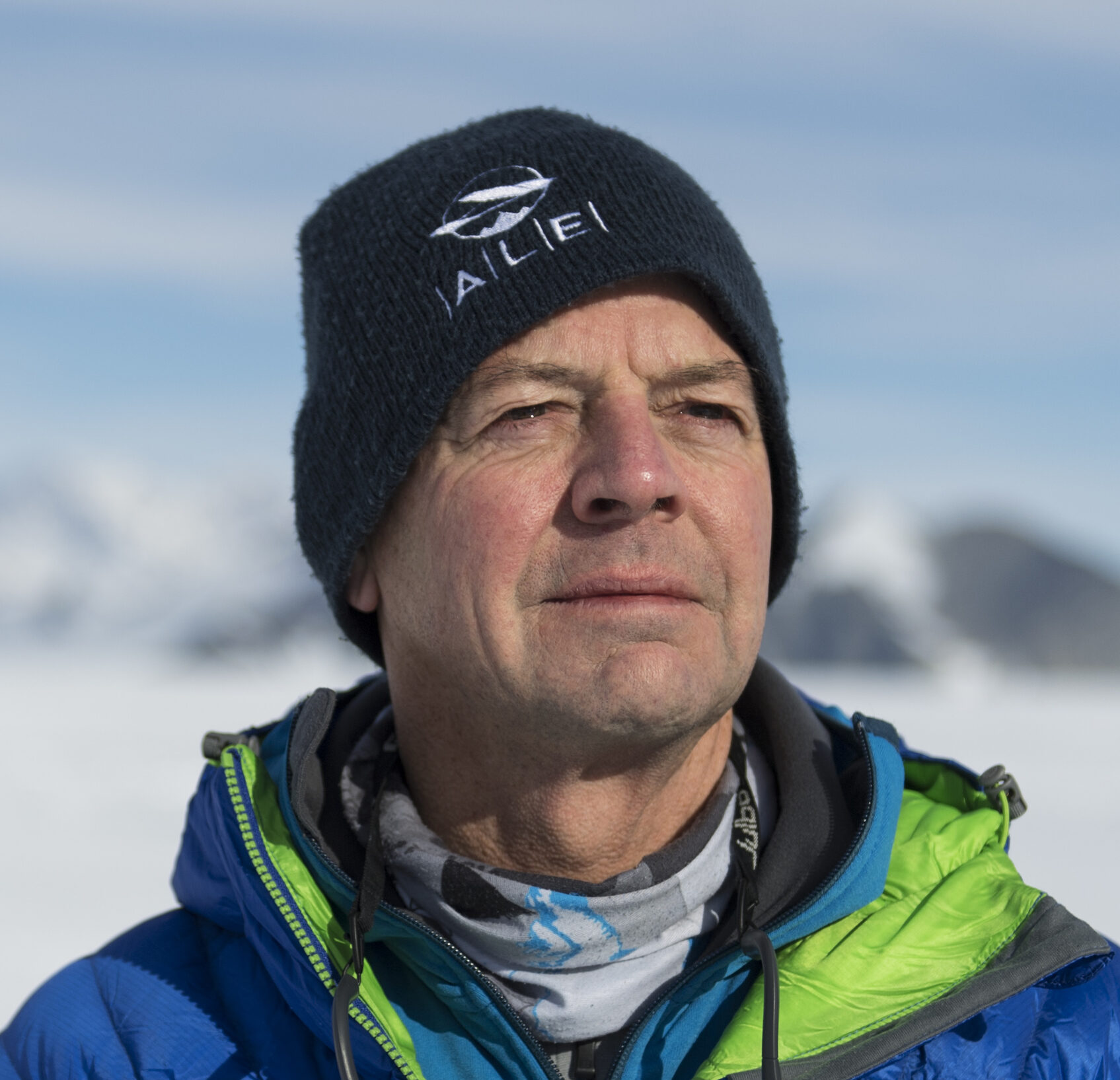 A man wearing a dark beanie and blue-green winter jacket stands outdoors in snowy, mountainous terrain near the South Pole, looking into the distance under bright daylight.