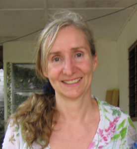 A woman with long, wavy blonde hair in a ponytail is smiling and wearing a floral-patterned top. Indoors, she stands near a window and wooden planks, sharing stories of her experiences with Antarctic Logistics and Expeditions.