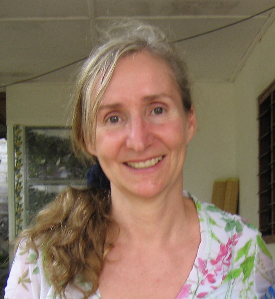 A woman with long, wavy blonde hair in a ponytail is smiling and wearing a floral-patterned top. Indoors, she stands near a window and wooden planks, sharing stories of her experiences with Antarctic Logistics and Expeditions.