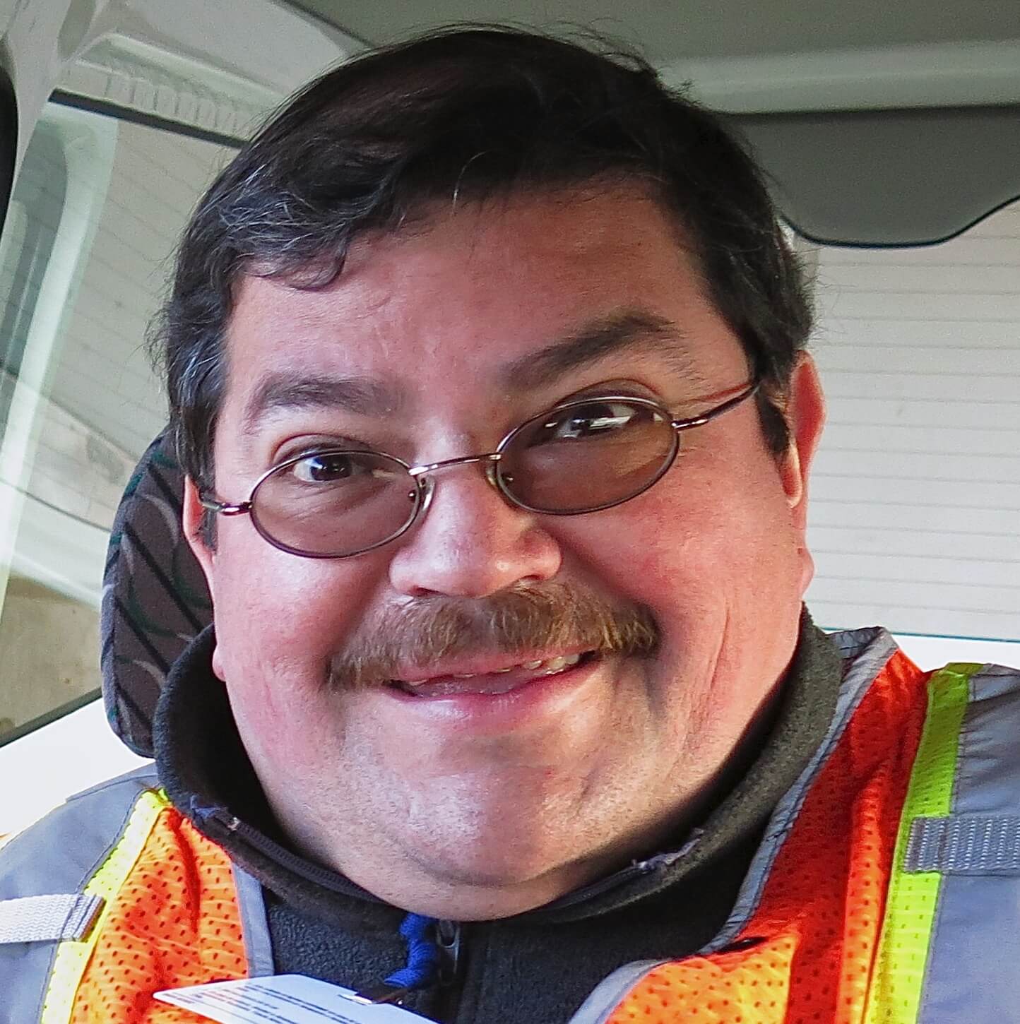 A man with glasses and a mustache smiles while wearing an orange safety vest and a dark jacket, sitting inside a vehicle, ready for his Antarctic Logistics and Expeditions adventure in Antarctica.